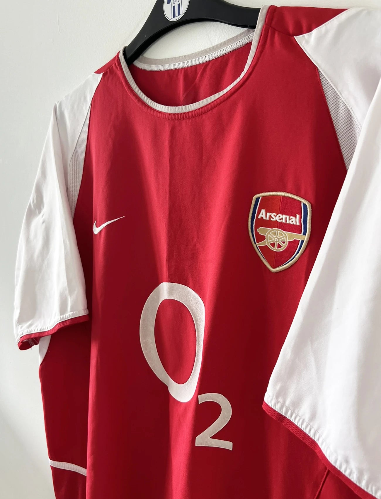 Maillot domicile Arsenal rouge et blanc de la saison 2002-2004. On peut retrouver l'équipementier nike et le sponsor O2. Il s'agit d'un maillot authentique d'époque comportant l'étiquette 184985