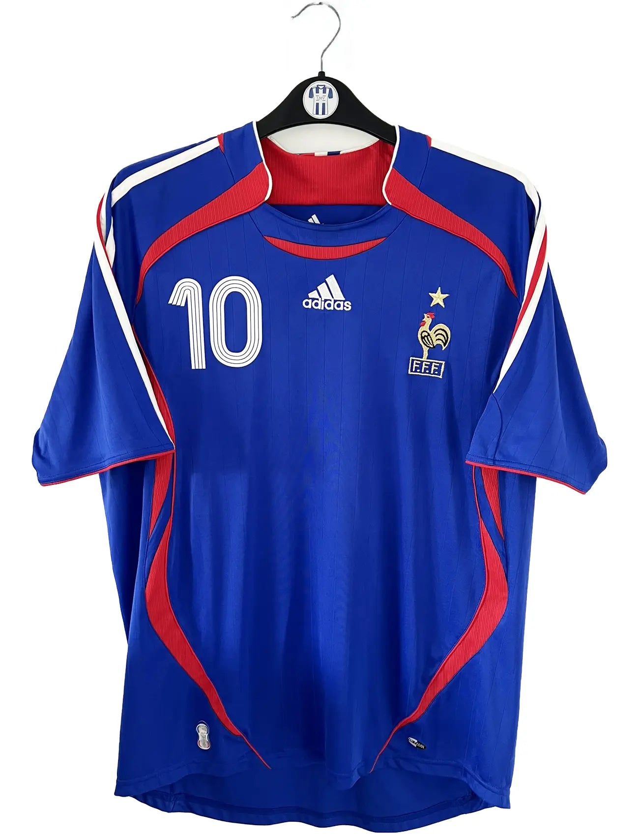 Maillot de foot vintage domicile bleu, blanc et rouge de l'équipe de france 2006. On peut retrouver l'équipementier adidas. Le maillot est floqué du numéro 10 Zinedine Zidane. Il s'agit d'un maillot authentique d'époque comportant l'étiquette 740126. Le maillot est un taille XL