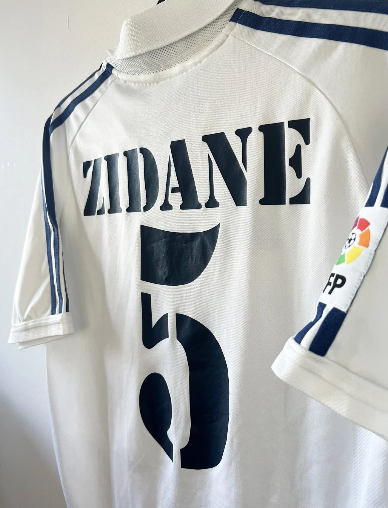 Maillot de foot vintage domicile du real madrid de la saison 2001-2002. Le maillot est de couleur blanc et noir. On peut retrouver l'équipementier adidas et le sponsor Real Madrid.com. Le maillot est floqué du numéro 5 Zinedine Zidane. Il s'agit d'un maillot authentique comportant les numéros 695856