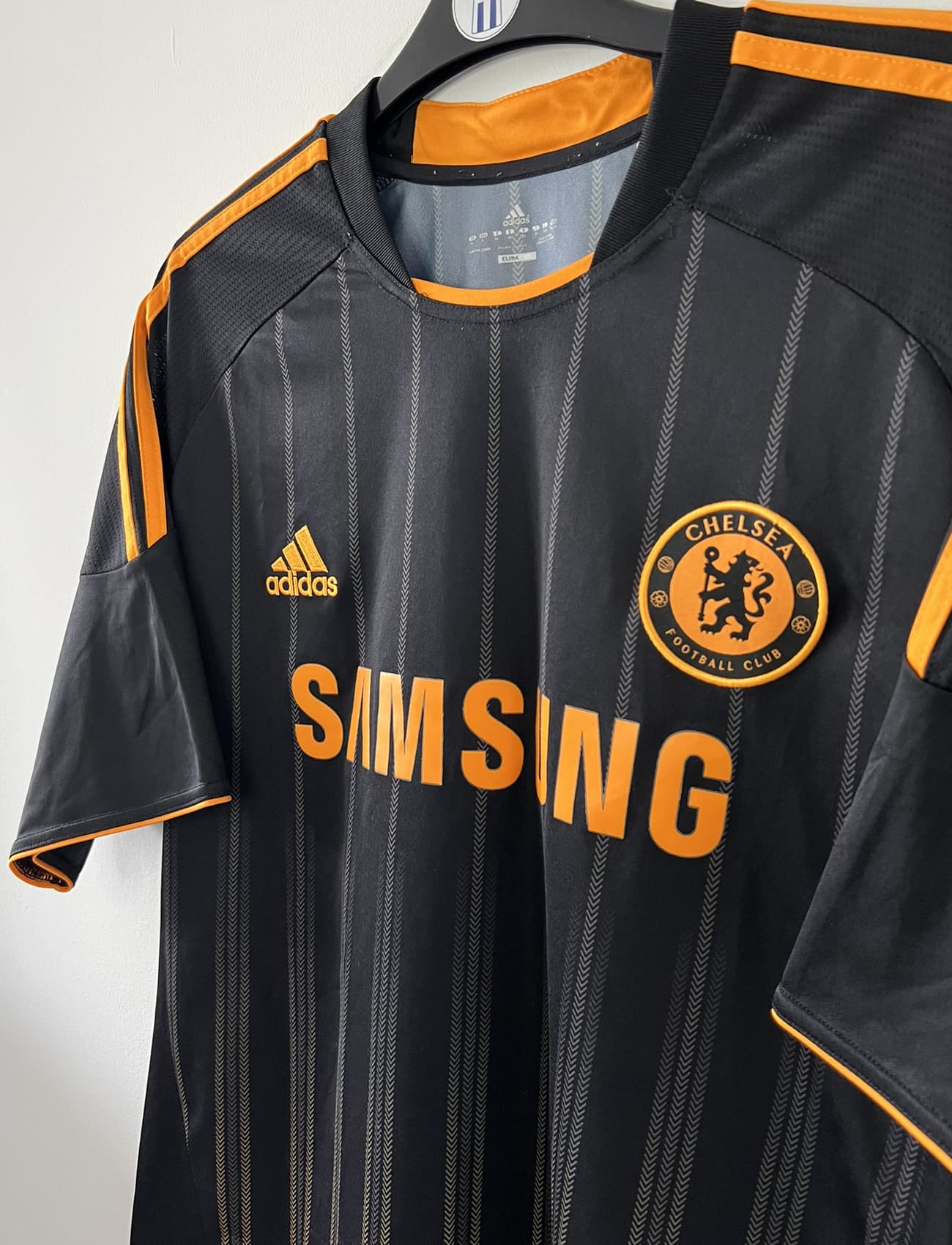 Maillot de foot vintage extérieur de chelsea de la saison 2010-2011. Le maillot est de couleur noir et orange. On peut retrouver l'équipementier adidas et le sponsor samsung. Le maillot est floqué du numéro 11 Didier Drogba. Il s'agit d'un maillot authentique comportant l'étiquette P00205