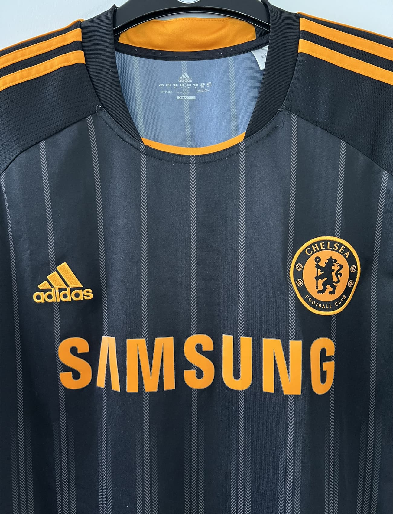 Maillot de foot vintage extérieur de chelsea de la saison 2010-2011. Le maillot est de couleur noir et orange. On peut retrouver l'équipementier adidas et le sponsor samsung. Le maillot est floqué du numéro 11 Didier Drogba. Il s'agit d'un maillot authentique comportant l'étiquette P00205