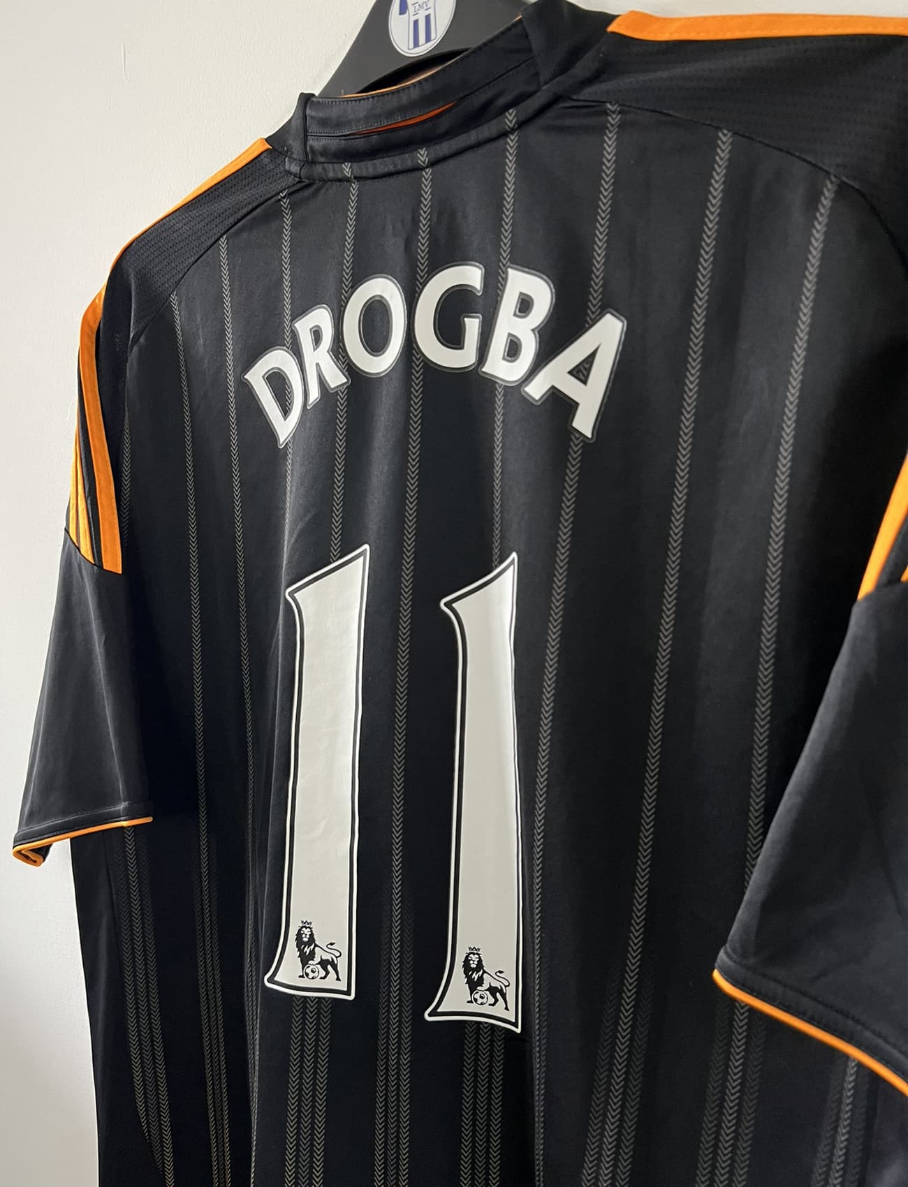 Maillot de foot vintage extérieur de chelsea de la saison 2010-2011. Le maillot est de couleur noir et orange. On peut retrouver l'équipementier adidas et le sponsor samsung. Le maillot est floqué du numéro 11 Didier Drogba. Il s'agit d'un maillot authentique comportant l'étiquette P00205