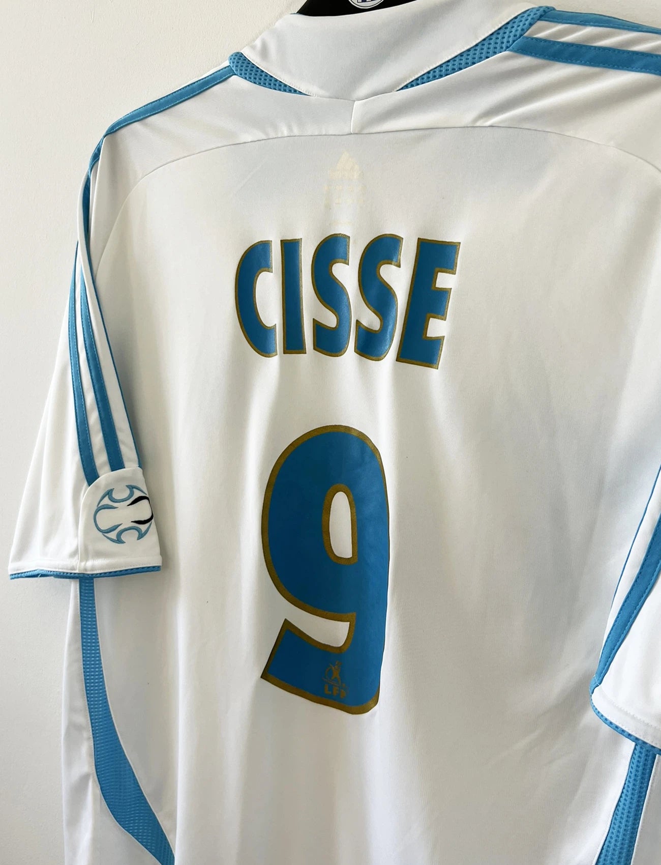 Maillot de foot vintage domicile de l'OM de la saison 2006/2007. Le maillot est de couleur blanc et bleu. On peut retrouver l'équipementier adidas et le sponsor Neuf. Le maillot est floqué du numéro 9 Djibril Cissé. Il s'agit d'un maillot authentique d'époque comportant les numéros 084929