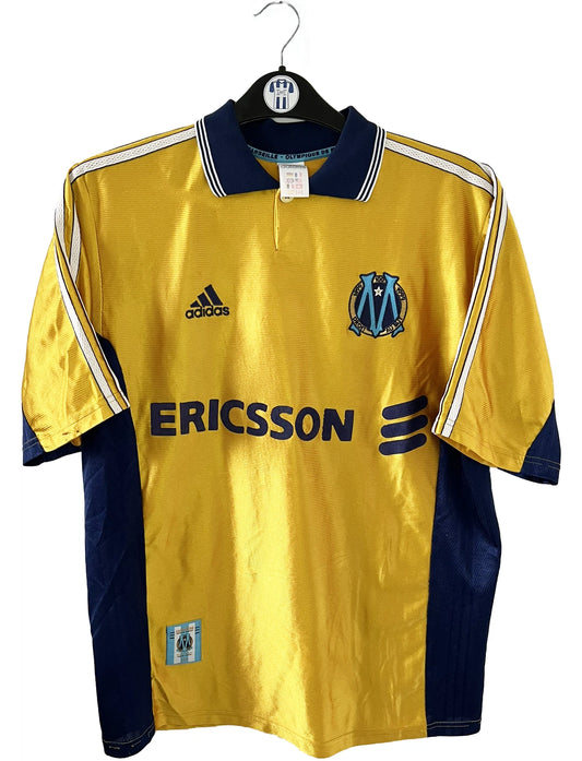 Maillot de foot vintage third de l'OM de la saison 1998-1999. Il s'agit du maillot édité pour le centenaire du club. Le maillot est de couleur dorée et bleu. On peut retrouver l'équipementier adidas et le sponsor ericsson. Il s'agit d'un maillot authentique d'époque.